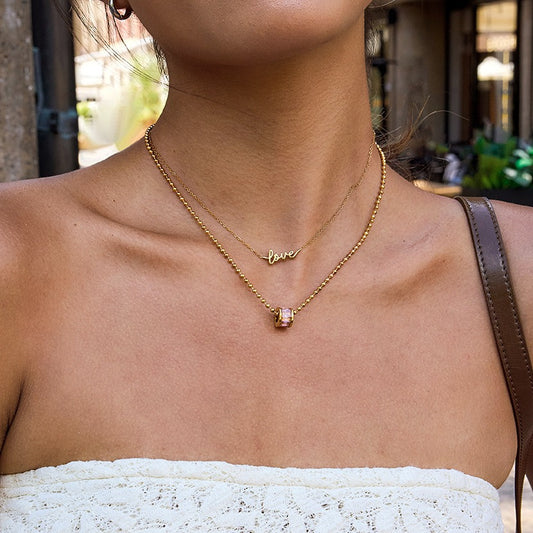 Gold layered necklaces with a "love" pendant and a small gemstone charm, worn on a woman's neck with a strapless top.